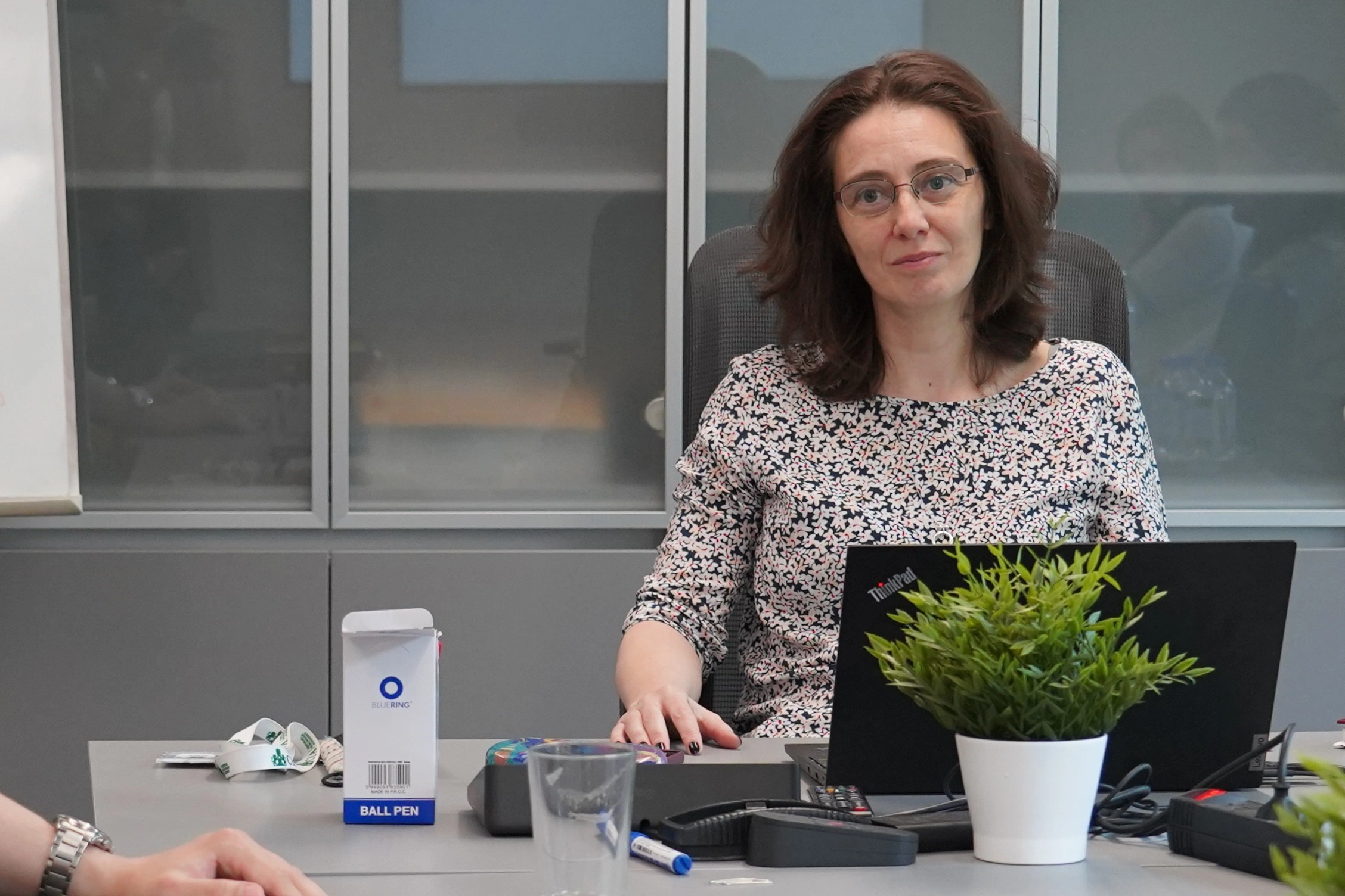 Nő ül a tárgyalóteremben laptop mögött, előtte pohár és jegyzetek.