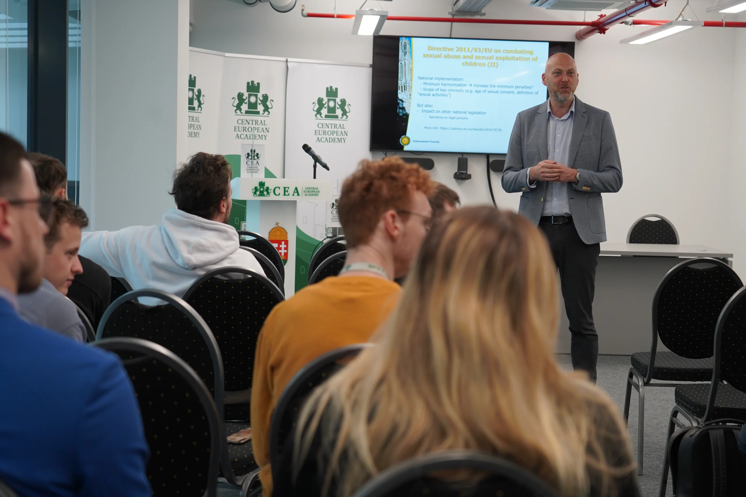 Male speaker giving a presentation to an audience at a Central European Academy event, with the screen showing the topic of the directive on combating the sexual abuse and exploitation of children.