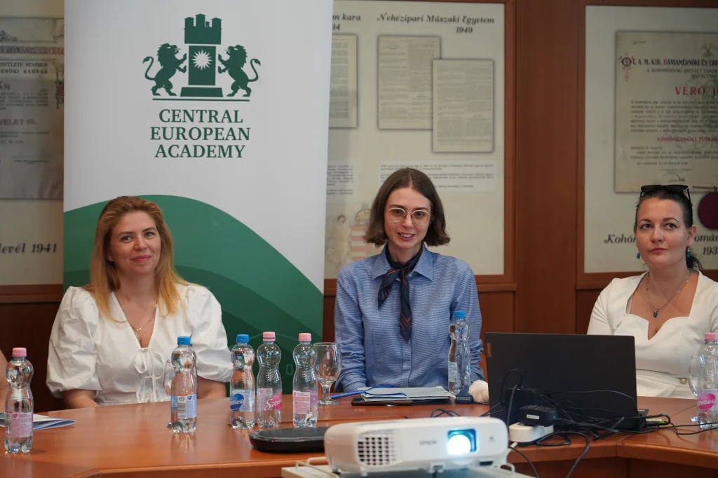 Három nő ül a tárgyalóasztalnál, mögöttük a Central European Academy logója.