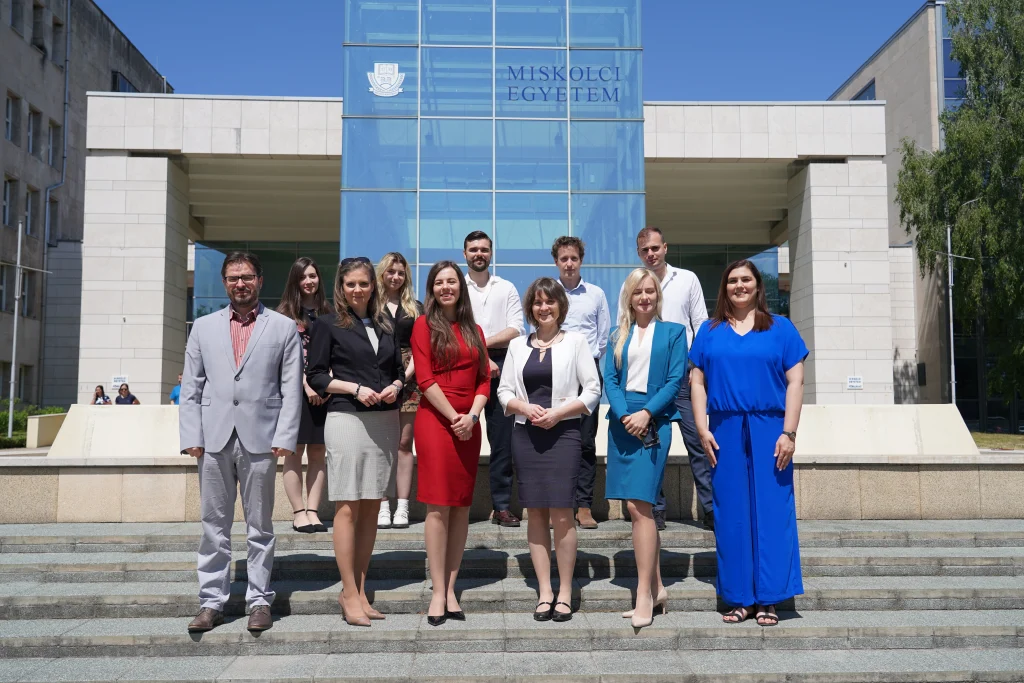 Csoportkép a Miskolci Egyetem épülete előtt, kék éggel a háttérben.
