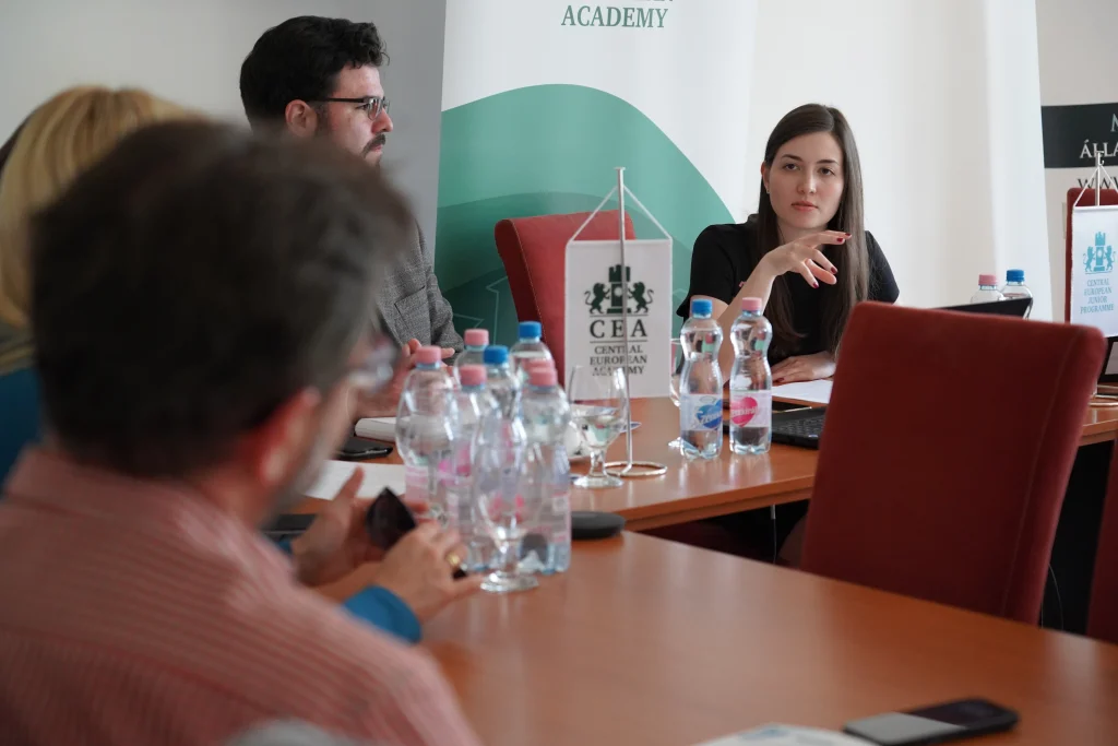 Résztvevők beszélgetnek egy tárgyalóban, az asztalon ásványvizes palackok és CEA zászló.